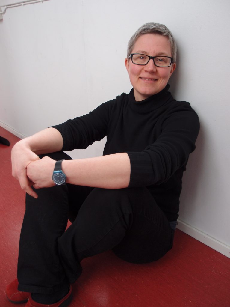Mustiin pukeutunut hymyilevä Rita istuu punaisella lattialla, kädet polvien ympärillä. Ritalla on lyhyet hiukset ja silmälasit.
