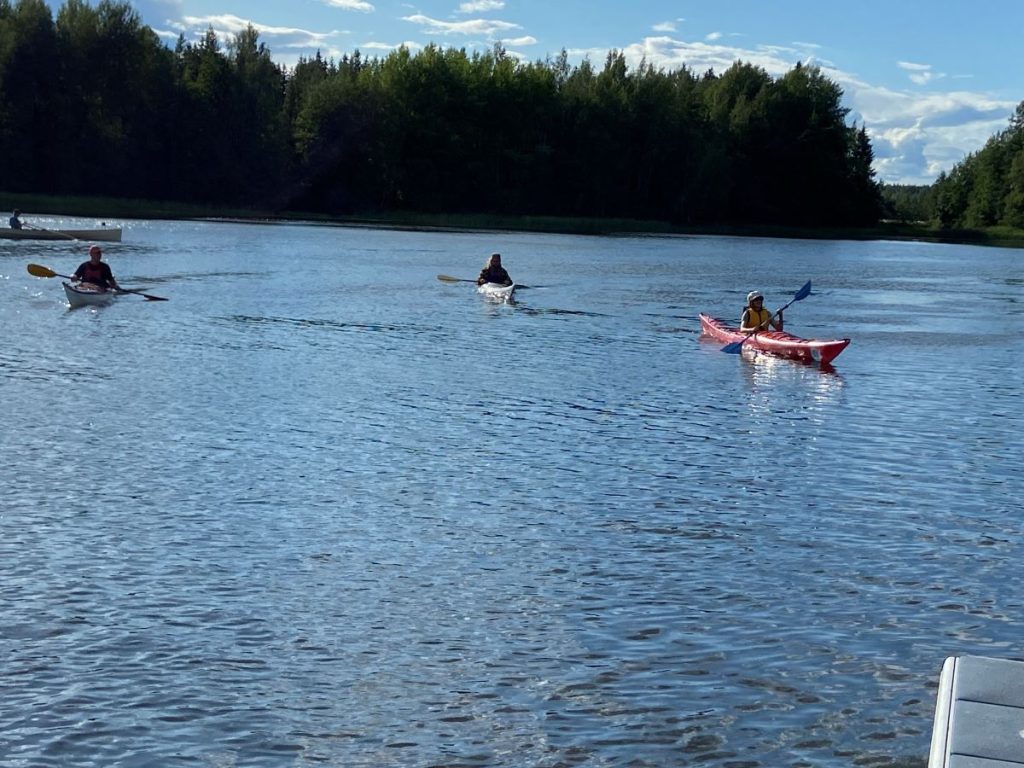 Kesäinen järvimaisema jossa 3 henkilöä kajakeilla melomassa ja 1 henkilö soutuveneessä.