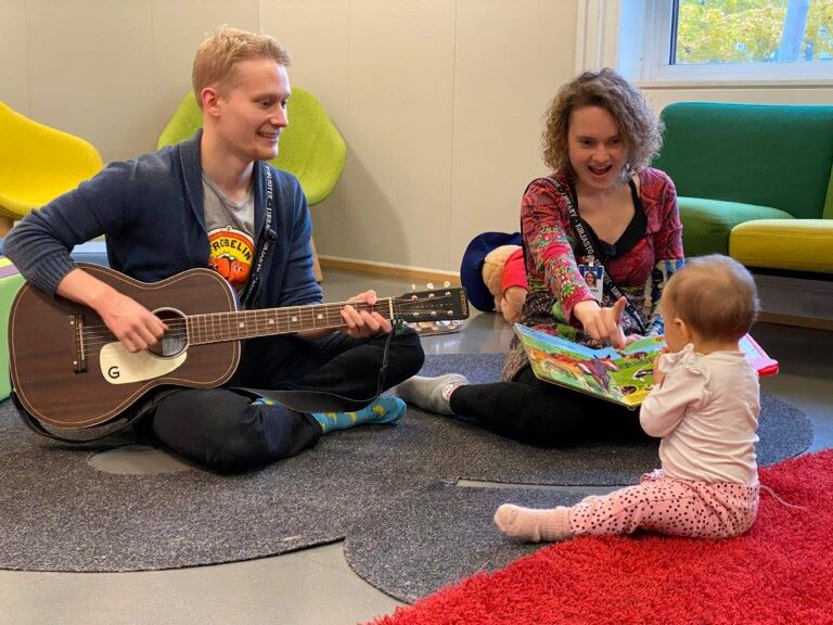 Mies soittaa kitaraa ja nainen lukee kuvakirjaa taaperolle. Kaikki istuu lattialla.