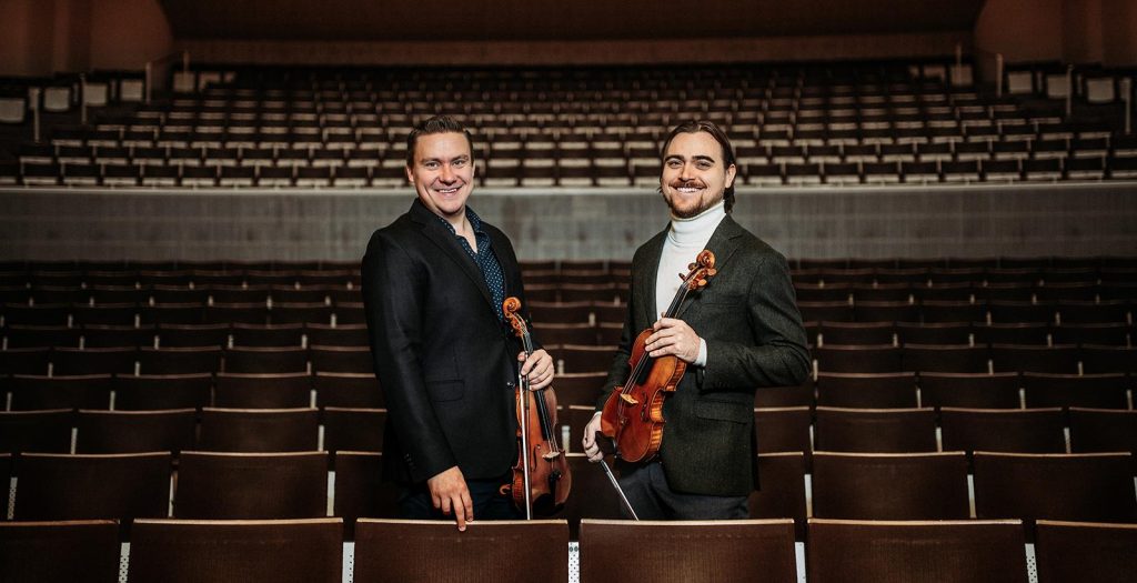 2 miestä seisoo konserttisalissa penkkirivien välissä. Heillä on viulut käsissään.