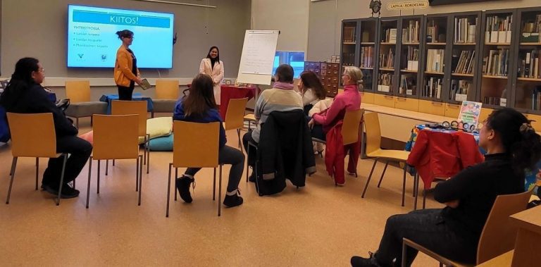 White assumedly woman and a brwn skinned and black haired assumedly woman in front of a small audience in a library.