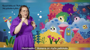 Liisa on pukeutunut violettiin mekkoon. Hänen takanaan on Meriyksisarvinen-kirjan värikäs aukeama, jossa ollaan meren pohjassa. Kalat ja muut meren elävät kurkkivat korallien välistä. Tekstitys: "huomaatko? Mukana on myös pallokala."