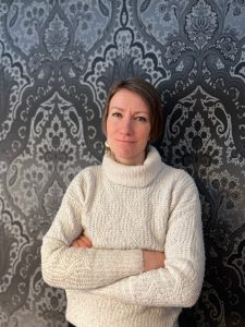 Salla seisoo harmaan kuviollisen tapetin edessä kädet puuskassa. Hänellä on vaaleanruskea polkkatukka ja valkoinen palmikko-pooloneule.