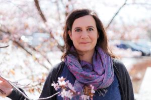 Ilaria tra i ciliegi in fiore. Ilaria ha la pelle chiara, i capelli castani e gli occhi grigi. Indossa una sciarpa viola intorno al collo.