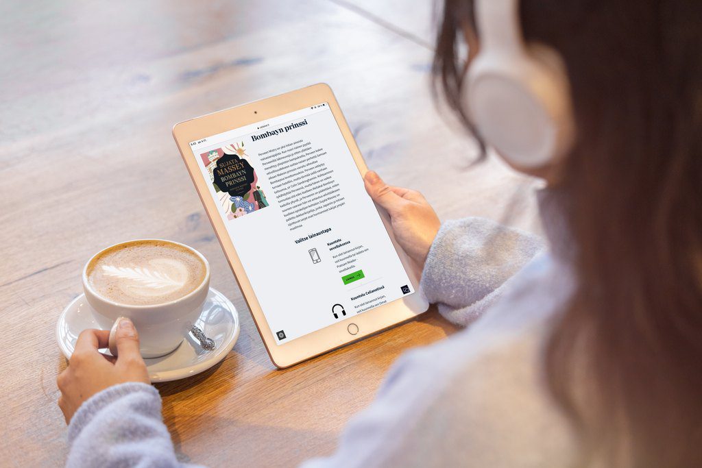 Takaviistosta kuvattu pitkätukkainen nuori lukee kuulokkeet päässä tabletilta kirjaa pöydän ääressä. Hän on tarttunut pöydällä olevaan kahvikuppiin.